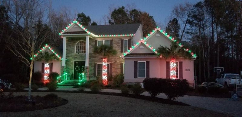 Two-story home with warm white roofline lights and red-and-green wreaths on the columns